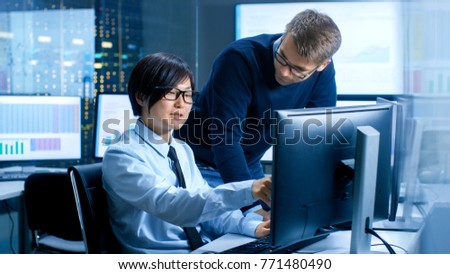 In the Data Mining Center Chief Statistician Consults His Young Assistant sits at His Workstation Surrounded with Monitors Displaying Data and Graphs.