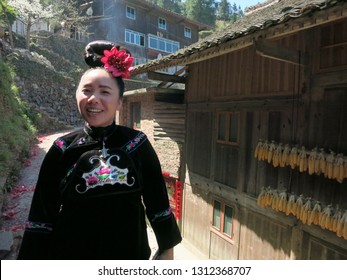 Datang, China - March 9, 2018: A smiling young miao woman in a traditional black shirt with embroidery and an artificial flower in her hair walking in the village street