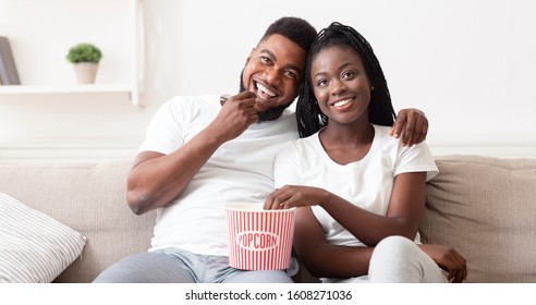 Date at home. Young afro couple embracing, eating popcorn and watching movie in living room, enjoying time together