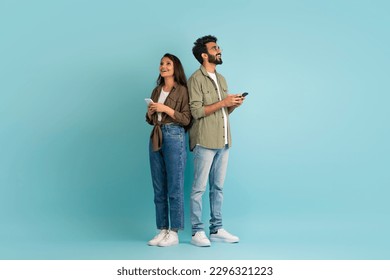 Dating mobile app concept. Stylish young eastern man and woman hipsters standing back to back, using smartphones on colorful studio background, chatting online, looking at copy space, full length
