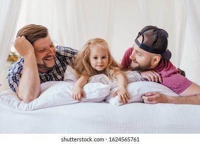 Daughter And Dads Resting On A Bed. Gay Parents Talking And Surrounding Their Child With Warm Atmosphere.