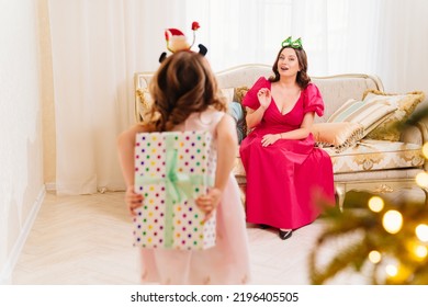 Daughter Hides The Gift Behind Her Back. A Happy Mom On The Couch By The Christmas Tree. Tradition Of Decorating House And Giving Gifts For The New Year. Surprises. Happy Childhood.