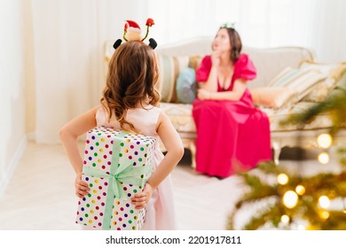 Daughter Hides The Gift Behind Her Back. A Happy Mom On The Couch By The Christmas Tree. Tradition Of Decorating House And Giving Gifts For The New Year. Surprises. Happy Childhood.