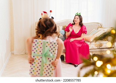 Daughter Hides The Gift Behind Her Back. A Happy Mom On The Couch By The Christmas Tree. Tradition Of Decorating House And Giving Gifts For The New Year. Surprises. Happy Childhood.