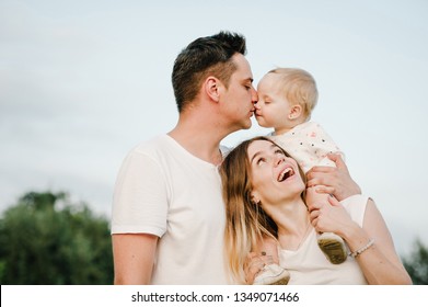 The Daughter Hugging Parents On Nature. Mom, Dad And Girl Toddler,  Walk In The Grass. Happy Young Family Spending Time Together, Outside, On Vacation, Outdoors. The Concept Of Family Holiday.