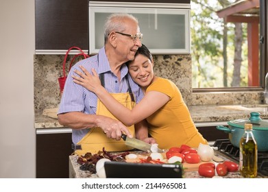 Daughter hugs her father tenderly in the kitchen while her father prepares food.