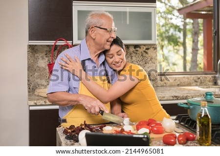 Daughter hugs her father tenderly in the kitchen while her father prepares food.