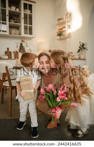 Daughter and little son kissing and hugging mom and make surprise for Mothers Day. Children give bouquet of tulip flowers. Childs gives present box congratulates surprised mom in kitchen. Happy family