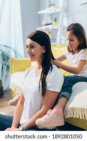 Daughter plaiting hair of smiling mother while sitting on couch at home on blurred background - Shutterstock ID 1892801050