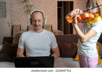 daughter play violin while Father making notes in notebook, looking at laptop. Young man holding child and working on PC. Dad doing homebased business. multi-tasking, freelance and fatherhood concept