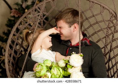 Daughter Plays With Dad. Babe Caught Father's Nose And Man's Eyes Were Closed. Funny Family Photo. Blonde Girl In A White Dress Sitting On Dad's Lap