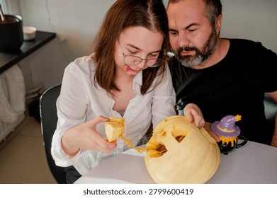 A daughter pulls stringy pulp and a carved piece from a jack-o-lantern while her father helps in a cozy kitchen. A warm autumn scene of a father and daughter preparing a jack-o-lantern for Halloween at home.