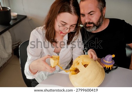 A daughter pulls stringy pulp and a carved piece from a jack-o-lantern while her father helps in a cozy kitchen. A warm autumn scene of a father and daughter preparing a jack-o-lantern for Halloween at home.