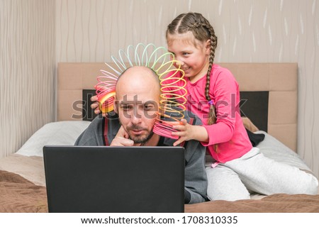 The daughter puts the slinky toy to the father's head. Family is quarantined at home. A little girl is making fun of dad.A child prevents a man from working on a laptop in his bedroom.