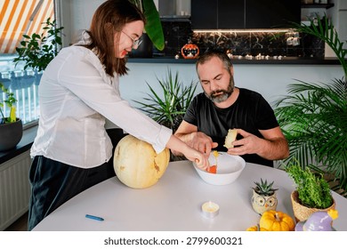 A daughter scoops stringy pulp from a pale pumpkin into a white bowl while her father helps at a festive table with mini pumpkins. A warm autumn scene of a father and daughter preparing a jack-o-lantern for Halloween at home.