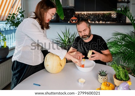 A daughter scoops stringy pulp from a pale pumpkin into a white bowl while her father helps at a festive table with mini pumpkins. A warm autumn scene of a father and daughter preparing a jack-o-lantern for Halloween at home.