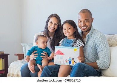 Daughter showing drawing happy family in new house  Cute infant looking at colorful drawing his sister  Happy proud multiethnic parents sitting and children sofa    looking at camera 