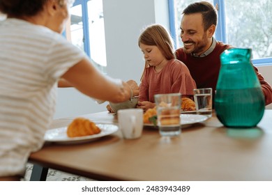 Daughter is sitting in her fathers lap, as mother pours the milk in her cereal bowl during family breakfast - Powered by Shutterstock