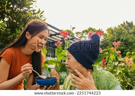 Daughter taking care health elderly mother who is sick with cancer sitting in wheelchair to eat soup to restore body and health but mother is bored of food doesn't want to eat bland food and can't eat Foto stock © 