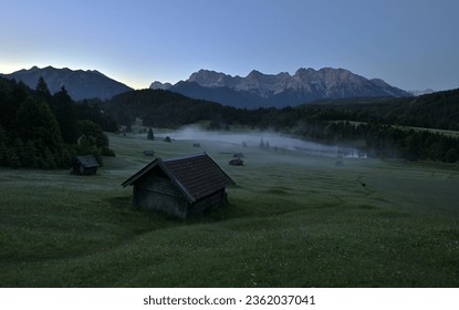 Dawn in The Bavarian Alps, Hay barns stand on a Mountain Meadow at Dusk, Alpine Panorama Early in The Morning, View of a Mountain Range and a Mountain Lake, An Idyllic Mountain Landscape,  - Powered by Shutterstock