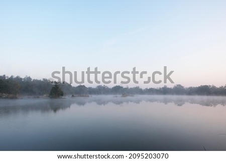 Dawn over the foggy lake. Beautiful dreamy view. Blue sky just before the sunrise and fog over water and trees with reflections on the river bank.