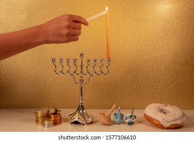 Day 1 of jewish religious holiday Hanukkah with child's hand lighting 1st candle in traditional chandelier menorah on golden glittering background with other traditional objects around