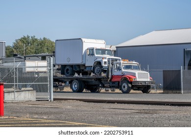 Day Cab Big Rig Semi Towing Truck Carry Another Truck With Box Trailer On Flat Bed Lifted Trailer Standing On The Industrial Warehouse Parking Lot