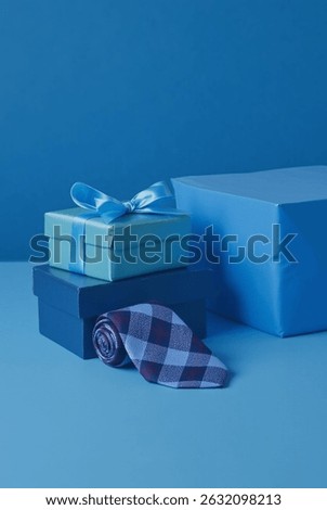 Father’s Day gift setup with two stacked gift boxes—one light blue with a satin ribbon, the other navy—accompanied by a rolled-up blue and burgundy checkered necktie. Ideal for Father's Day, birthdays
