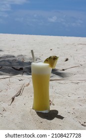 A day in the Maldives, a glass of freshly squeezed pineapple juice with white foam, an inserted straw and a piece of pineapple on the edge, standing on the white sand on the Indian Ocean
