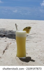A day in the Maldives, a glass of freshly squeezed pineapple juice with white foam, an inserted straw and a piece of pineapple on the edge, standing on the white sand on the Indian Ocean