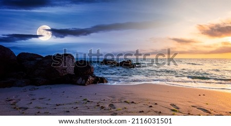 day and night time change concept above huge stones on the sandy beach at twilight. wonderful velvet season vacation. clouds on the blue sky with sun and moon above horizon. waves washing the shore
