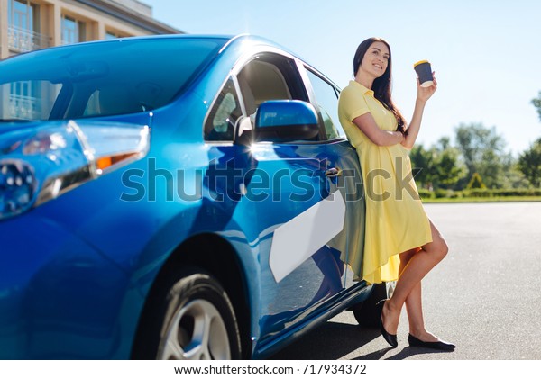 Day off work. Delighted\
beautiful happy woman standing near her car and resting while\
drinking coffee