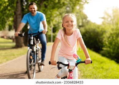 Day in the park. Happy family of caucasian father and girl child riding bicycles outdoors in the summer, smiling at camera. Dad and daughter's park adventure - Powered by Shutterstock