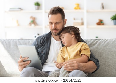 Day Rest. Young Peaceful Man Reading News Online On Digital Tablet And Carrying His Sleeping Little Son On Hands, Resting On Couch At Home