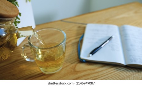 Day Summing In Bullet Journal Planner In Evening Light Room. Close-up, Herbal Tea Poured Into Cup Of Glass Teapot. Open Paper Diary On Wooden Table Lies With Notes, Pen Lies On Pages. Year Sum Results