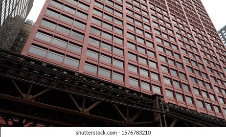 Day Time Exterior Establishing Shot Of Generic Large Red Skyscraper In Chicago. Loop Train Tracks Pass By Street Level Outside Building Facade For Commute To Corporate Headquarters