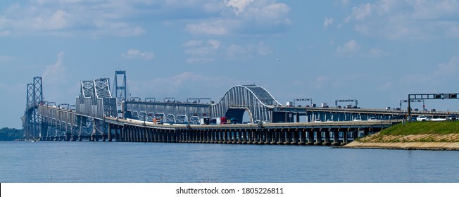A day time wide angle image showing the rush hour traffic on Chesapeake Bay Bridge. It features detailed view of the bridge with columns and suspensions as well as the boats passing under.