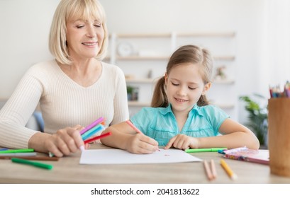 Daycare Activity, Babysitting Concept. Happy Mature Older Woman Sitting At Desk With Smiling Adorable Preschool Little Kid Girl, Enjoying Drawing Together In Paper Album, Using Coloful Markers