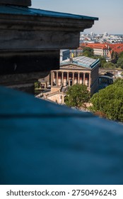 Daylight view from an elevated point shows the Alte Nationalgalerie portico and rooftop sculpture through blurred ledges, with treetops, red roofs, and skyline receding.