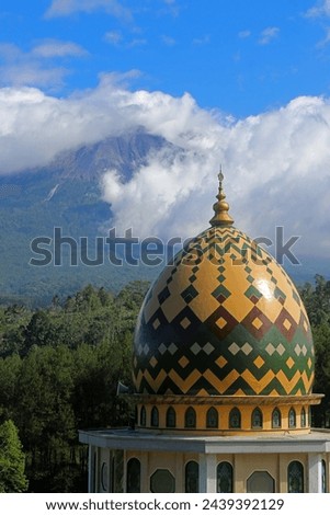 Daylight view of a village with a mosque dome in the foreground and cloudy Mount Semeru in the background. Thematic images for Ramadan and Eid.