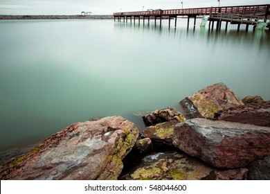 Daytime Long Exposure Winter Picture Of Stones In Foreground And Green Water Of Lake Erie And Dock In Background In Buffalo, New York