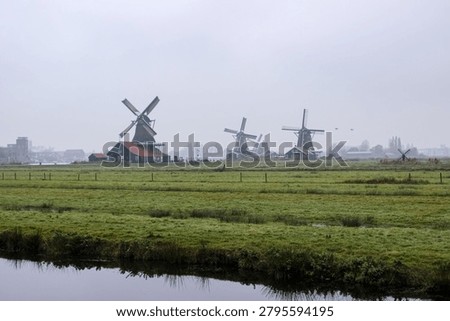 A daytime panoramic view capturing historic wooden windmills across green pastures and a calm canal on a misty, overcast day in the Netherlands.