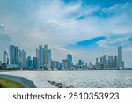 A daytime view of Panama City’s modern skyline seen from a waterfront promenade, with tall skyscrapers reflecting in the calm water and a palm tree adding a tropical touch to the urban landscape.