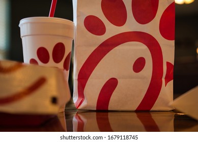 Dayton, OH- Mar 16, 2020; Chick Fil A Bag With Other Food Items In Background. Boxes And Bags Of Trash Following Meal. Reflection On Wooden Table