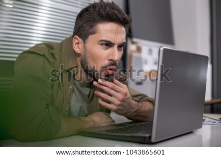 Dazed bearded guy sitting at table in his workplace and looking at notebook screen with astonishment