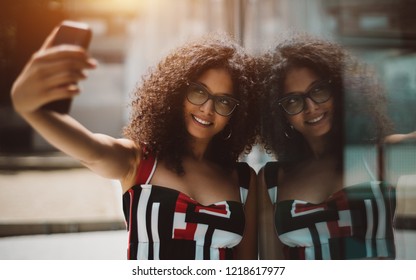 Dazzling hipster girl in eyeglasses and with bulky curly hair is taking a selfie using her smartphone while leaning against the glass wall outdoors which fully reflects her and smiling - Powered by Shutterstock