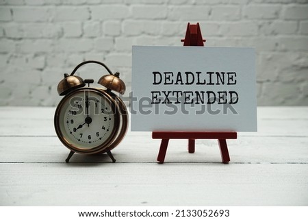 Deadline Extended text and alarm clock on white brick wall and wooden background