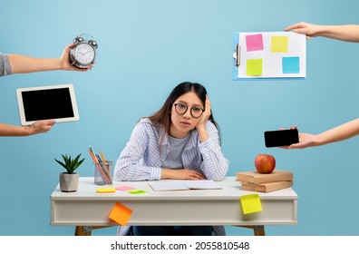 Deadline and multitask concept. Tired asian lady stressed by a lot of work, sitting at the desk over blue background and looking at camera. Hands with tablet, watch, tasks and smartphone