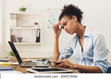 Deadline stress concept - sad african-american business woman sitting at desktop in office, working on laptop and holding hand on head. Hard working day, copy space
