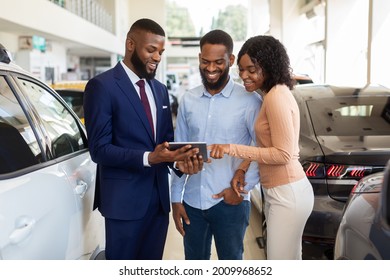 Dealership Center Manager Showing Vehicle Characteristics On Digital Tablet To Young Black Couple, Cheerful African American Spouses Purchasing New Vehicle In Auto Showroom, Closeup Shot